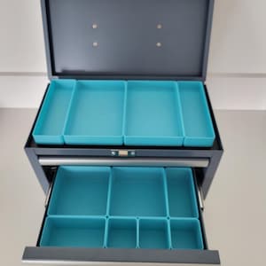 May include: A gray metal storage chest with a hinged lid and one pull-out drawer. The interior features multiple removable bright turquoise plastic organizer trays of varying rectangular sizes, designed to keep small items separated and organized.