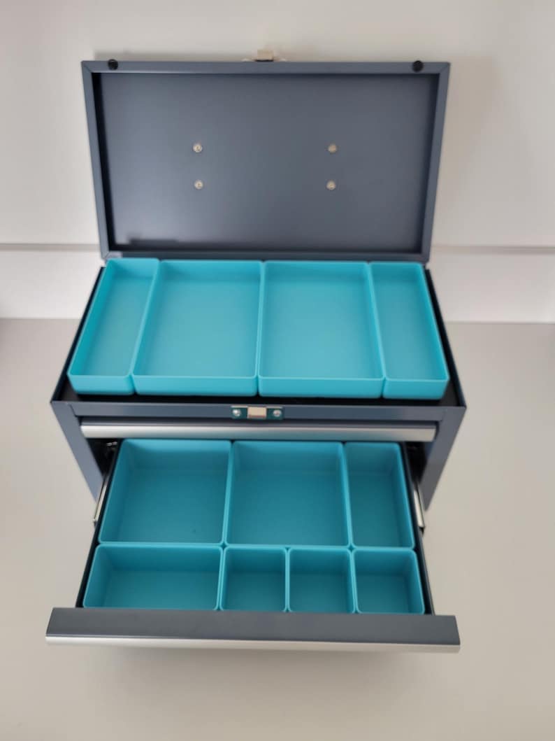 May include: A gray metal storage chest with a hinged lid and one pull-out drawer. The interior features multiple removable bright turquoise plastic organizer trays of varying rectangular sizes, designed to keep small items separated and organized.