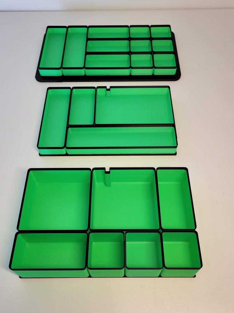 May include: Three rectangular drawer organizers featuring a bright green interior and a black exterior frame. Each organizer contains multiple compartments of varying sizes, designed with straight walls and flat bases for modular storage organization.