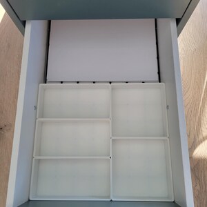 May include: An open gray drawer containing a white rectangular paper sheet and four white plastic storage trays. The trays are arranged in a grid pattern, with two smaller rectangular trays on the left and two larger square trays on the right.