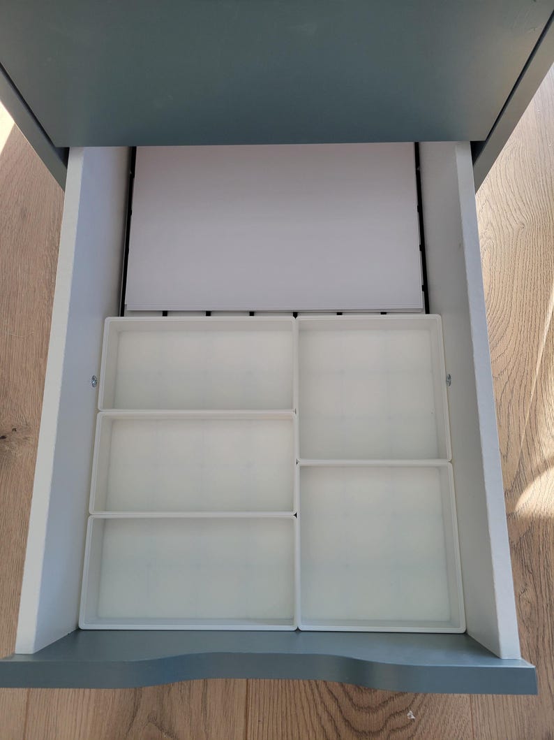 May include: An open gray drawer containing a white rectangular paper sheet and four white plastic storage trays. The trays are arranged in a grid pattern, with two smaller rectangular trays on the left and two larger square trays on the right.