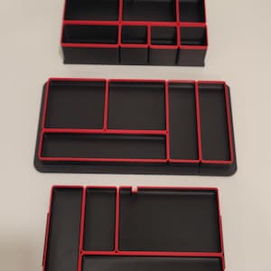 May include: Three black rectangular desk organizers with vibrant red trim. Each organizer features multiple compartments of varying sizes for storage. The design includes clean lines and a structured layout, providing organized sections for small items.