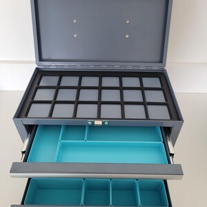 May include: A gray metal storage chest with a hinged lid and two pull-out drawers. The top section features a black foam insert with fifteen square compartments. The two lower drawers contain bright turquoise plastic organizers for sorting items.