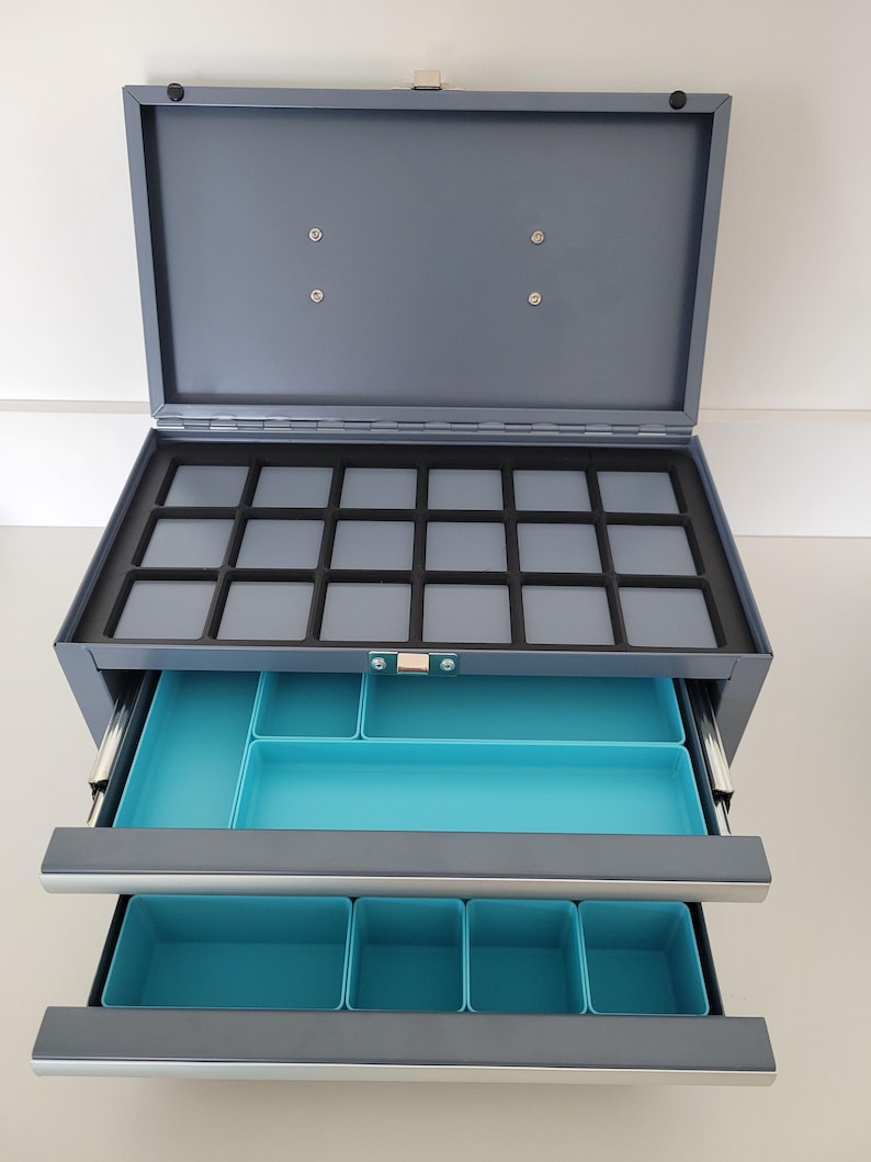 May include: A gray metal storage chest with a hinged lid and two pull-out drawers. The top section features a black foam insert with fifteen square compartments. The two lower drawers contain bright turquoise plastic organizers for sorting items.