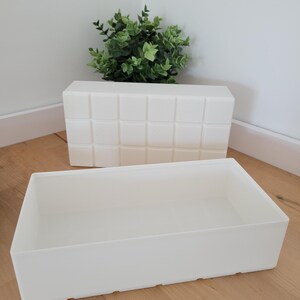 May include: Two white rectangular storage containers. One has a grid pattern on the front. The other is a simple, open box. A small green plant sits behind the grid-patterned container.