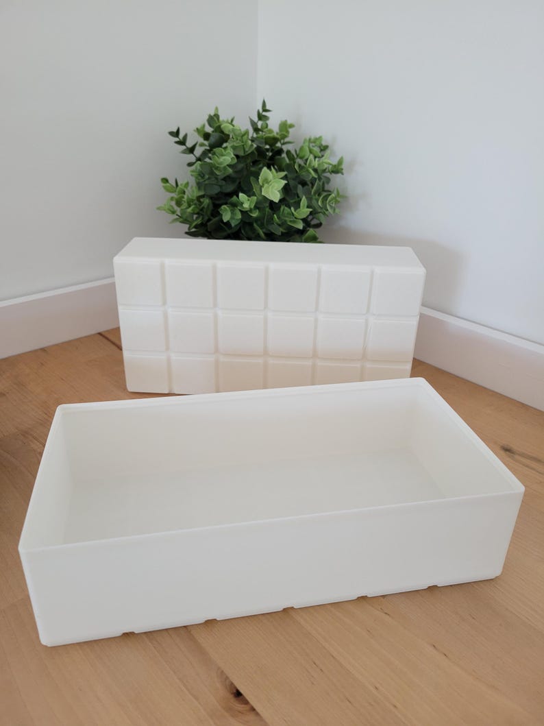 May include: Two white rectangular storage containers. One has a grid pattern on the front. The other is a simple, open box. A small green plant sits behind the grid-patterned container.