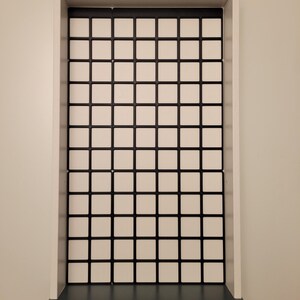 May include: A rectangular white wall-mounted display shelf featuring a grid-patterned back panel with black dividers and white square inserts. The unit has a solid white frame and a dark gray base shelf with a curved front edge for storage.
