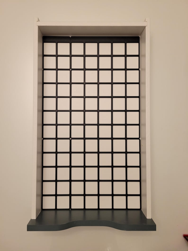 May include: A rectangular white wall-mounted display shelf featuring a grid-patterned back panel with black dividers and white square inserts. The unit has a solid white frame and a dark gray base shelf with a curved front edge for storage.