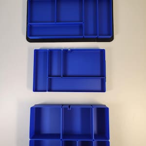 May include: Three rectangular blue plastic drawer organizers with various compartment configurations. Each tray features multiple open sections of different sizes for sorting items, set against a plain background for clear visibility.