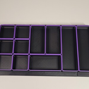 May include: A rectangular drawer organizer tray featuring a black base and multiple black compartments with purple rims. The organizer includes various sizes of square and rectangular sections arranged in a grid pattern for storage and organization.