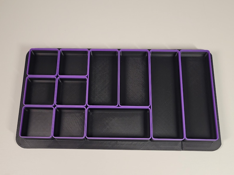 May include: A rectangular drawer organizer tray featuring a black base and multiple black compartments with purple rims. The organizer includes various sizes of square and rectangular sections arranged in a grid pattern for storage and organization.