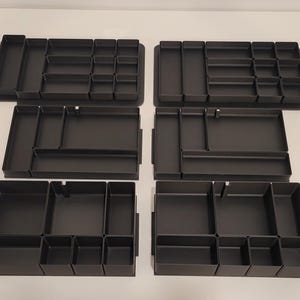 May include: A set of six black drawer organizers arranged in two columns. Each organizer features multiple rectangular and square compartments of varying sizes, designed with solid walls and flat bases for sorting small items within a drawer.