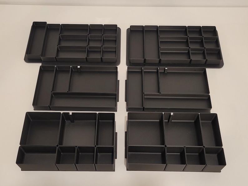 May include: A set of six black drawer organizers arranged in two columns. Each organizer features multiple rectangular and square compartments of varying sizes, designed with solid walls and flat bases for sorting small items within a drawer.