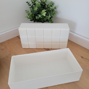 May include: Two white rectangular storage containers. One is open, revealing an empty interior. The other is closed, with a grid-like pattern on the front. A small green plant sits in the background. The containers are on a wooden surface. Ideal for home storage.