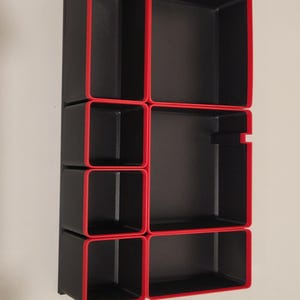 May include: A rectangular black storage organizer tray featuring multiple compartments of varying sizes, each defined by a bold red border along the top edges. The unit is structured with a grid-like layout, providing several open sections for sorting items.
