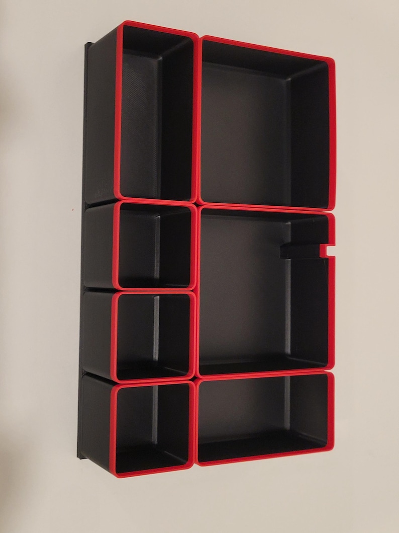 May include: A rectangular black storage organizer tray featuring multiple compartments of varying sizes, each defined by a bold red border along the top edges. The unit is structured with a grid-like layout, providing several open sections for sorting items.