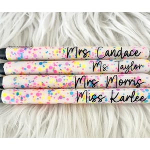 May include: Five white pens with colorful confetti glitter on the surface. The pens are labeled with the names "Mrs. Candace", "Ms. Taylor", "Mrs. Morris", and "Miss Karlee".