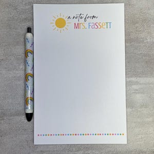 May include: White notepad with a rainbow dotted border and the text "a note from Mrs. Fassett" in colorful lettering. A pen with a rainbow and glitter design rests on the left side of the notepad.