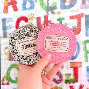 May include: Two round composition book-themed coasters. One is black with silver glitter, the other is pink with glitter. Each coaster has a label that reads "Notes" and dimensions of 9 ¾ x 7 ½ inches.
