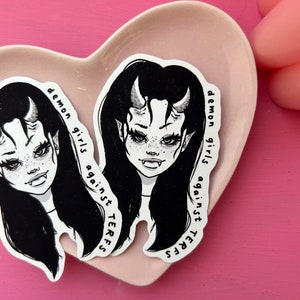 May include: Two stickers with a black and white illustration of a demon girl with horns, long black hair, and fangs. The text "demon girls against TERFS" is printed on the side of each sticker. The stickers are on a heart-shaped plate.