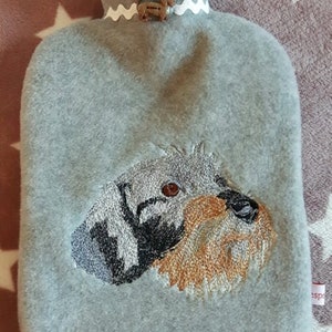 Könnte beinhalten: Eine graue Wärmflasche mit einem gestickten Hundegesicht. Die Flasche hat ein dekoratives weißes Band mit einem kleinen knopfförmigen Hund. Das Hundegesicht ist mit braunem, schwarzem und grauem Faden detailliert.