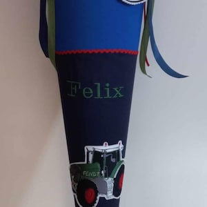 Könnte beinhalten: Ein blauer und marineblauer Schultüte mit rotem Besatz. Die Tüte hat ein Traktor-Design und den Namen "Felix" aufgestickt. Ein weißer Kreis mit dem Text "Schulkind 2023" ist an der Tüte befestigt.