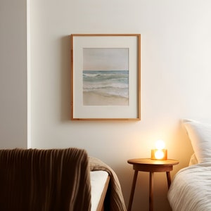 K&ouml;nnte beinhalten: Ein Schlafzimmer-Interieur mit einem Bett, einem Nachttisch und einem gerahmten Aquarellbild einer Meereslandschaft. Das Bild h&auml;ngt an einer wei&szlig;en Wand, und der Nachttisch hat eine Lampe mit einem wei&szlig;en Lampenschirm.
