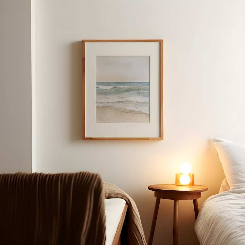 K&ouml;nnte beinhalten: Ein Schlafzimmer-Interieur mit einem Bett, einem Nachttisch und einem gerahmten Aquarellbild einer Meereslandschaft. Das Bild h&auml;ngt an einer wei&szlig;en Wand, und der Nachttisch hat eine Lampe mit einem wei&szlig;en Lampenschirm.