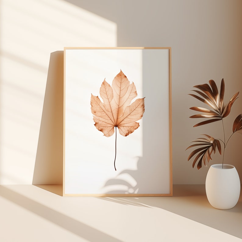 May include: A framed art print featuring a detailed, sepia-toned leaf against a white background. The leaf's intricate veins and edges are visible. The frame is a light wood color. A white vase with decorative plants is to the right.