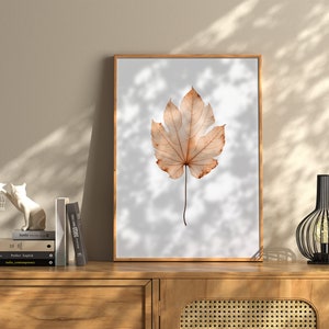 May include: A framed print featuring a detailed watercolor illustration of a single, large, dried leaf in shades of brown and tan. The leaf is centered against a light gray background, encased in a wooden frame. The artwork is displayed on a wooden cabinet.