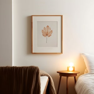 May include: A framed print featuring a watercolor illustration of a single, dried leaf in shades of brown and beige. The artwork is set against a cream-colored background and framed in a light wood frame. The print hangs on a white wall.