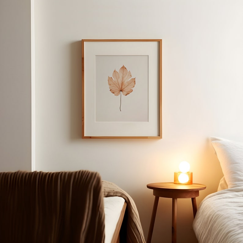 May include: A framed print featuring a watercolor illustration of a single, dried leaf in shades of brown and beige. The artwork is set against a cream-colored background and framed in a light wood frame. The print hangs on a white wall.