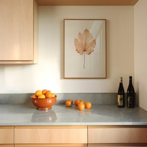 May include: A kitchen scene featuring a framed botanical print of a leaf, a terracotta bowl filled with oranges, and two dark wine bottles. The countertop is gray, and the cabinets are light wood.