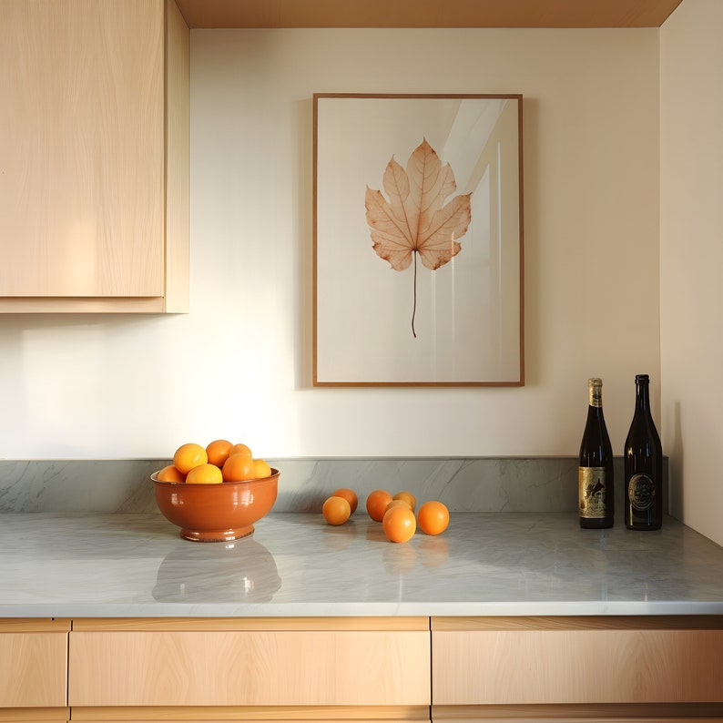 May include: A kitchen scene featuring a framed botanical print of a leaf, a terracotta bowl filled with oranges, and two dark wine bottles. The countertop is gray, and the cabinets are light wood.