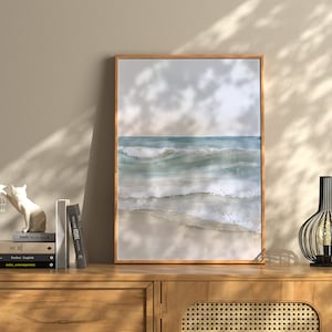 K&ouml;nnte beinhalten: Ein gerahmtes Aquarellbild eines Meeresbildes mit blauen und gr&uuml;nen Wellen, die an einem Sandstrand brechen. Das Bild h&auml;ngt an einer Wand &uuml;ber einer Kommode aus Holz mit einer Lampe und einer wei&szlig;en Katzenfigur.