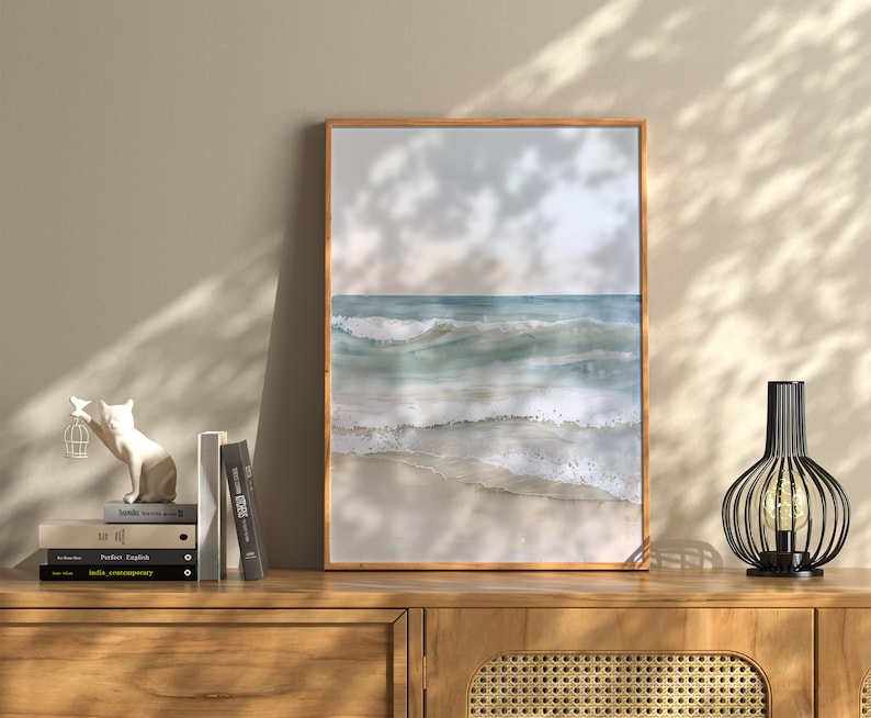 K&ouml;nnte beinhalten: Ein gerahmtes Aquarellbild eines Meeresbildes mit blauen und gr&uuml;nen Wellen, die an einem Sandstrand brechen. Das Bild h&auml;ngt an einer Wand &uuml;ber einer Kommode aus Holz mit einer Lampe und einer wei&szlig;en Katzenfigur.
