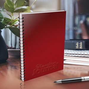 May include: A red spiral bound journal with the words "Transform Journal" embossed on the cover. The journal is sitting on a wooden table next to a stack of books and a pen.