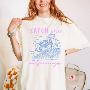 Pode incluir: Camiseta creme com um desenho de uma tartaruga surfando em uma onda azul. O texto "CATCH WAVES not feelings" est&aacute; em rosa e azul. A camiseta tem um corte folgado.