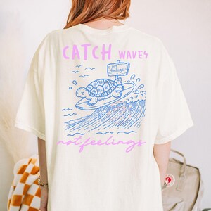 Pode incluir: Camiseta creme com um gr&aacute;fico azul e rosa. O design apresenta uma tartaruga surfando, ondas e o texto "Catch Waves not feelings". A camisa tem um corte folgado.