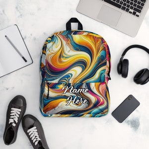 May include: A colourful backpack with an abstract pattern of swirling yellow, blue, pink, and white. The text "Name Here" is printed on the front. The backpack is next to a laptop, headphones, a phone, a notebook, a pencil, and black trainers.