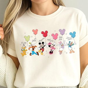 K&ouml;nnte beinhalten: Elfenbeinfarbenes T-Shirt mit einem farbenfrohen Aufdruck von Disney-Figuren, die Mickey Mouse-f&ouml;rmige Ballons halten. Die Figuren sind Pluto, Goofy, Mickey Mouse, Minnie Mouse, Daisy Duck und Donald Duck. Das Wort "Ivory" ist unten aufgedruckt.