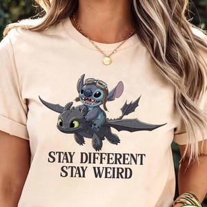 May include: Cream-coloured t-shirt with a cartoon image of Stitch riding a dragon, and the words "STAY DIFFERENT STAY WEIRD". The design is printed in black. The shirt has short sleeves and is made of a soft material.