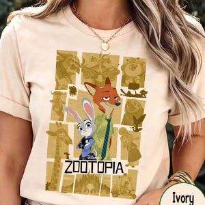 May include: Ivory-colored t-shirt featuring a graphic from the Disney movie Zootopia. The design includes characters Nick Wilde and Judy Hopps, with the movie title below. The shirt has short sleeves and a crew neck.
