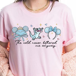 Op de afbeelding: Lichtroze T-shirt met een afbeelding van drie cartoon elementen: een Mickey Mouse oor hoofdband met een kroon, een hagedis en een Minnie Mouse oor hoofdband met een strik. De tekst "The cold never bothered me anyway" staat eronder.