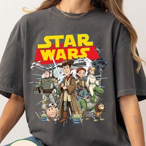 May include: Dark grey Comfort Colors t-shirt with a cartoon graphic of Star Wars characters. The Star Wars logo is in yellow and red. The t-shirt has a relaxed fit.