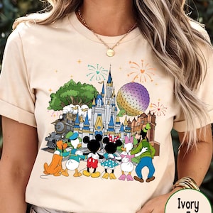 Peut inclure: T-shirt de couleur ivoire avec un motif color&eacute; de personnages et de monuments Disney, dont un ch&acirc;teau, un train et Spaceship Earth. Le motif comprend Mickey et Minnie Mouse, Donald Duck et Dingo. Le t-shirt a des manches courtes et un col rond.