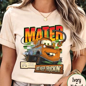 Puede incluir: Camiseta de color marfil con un dise&ntilde;o de Mate de la pel&iacute;cula Cars. El dise&ntilde;o incluye la palabra "MATER" en negrita, una ilustraci&oacute;n de un cami&oacute;n de remolque y la frase "JUST KEEP TRUCKIN'". La camiseta tambi&eacute;n tiene un logotipo de "Towing Salvage".