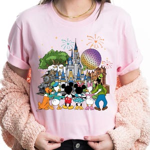 Peut inclure: T-shirt rose clair avec un motif color&eacute; de personnages Disney, dont Mickey et Minnie Mouse, Donald et Daisy Duck, et Dingo, devant un ch&acirc;teau, un train et une grande sph&egrave;re. Des feux d'artifice sont en arri&egrave;re-plan.