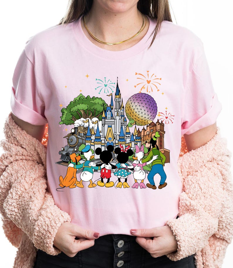 Peut inclure: T-shirt rose clair avec un motif color&eacute; de personnages Disney, dont Mickey et Minnie Mouse, Donald et Daisy Duck, et Dingo, devant un ch&acirc;teau, un train et une grande sph&egrave;re. Des feux d'artifice sont en arri&egrave;re-plan.