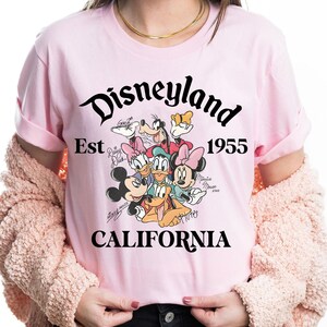 May include: A light pink t-shirt featuring the Disneyland logo with the text "Est 1955 CALIFORNIA". The shirt has a graphic of Disney characters including Mickey Mouse, Minnie Mouse, Donald Duck, and Goofy.